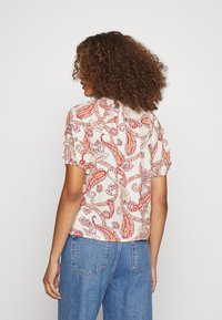 Blusa de mangas curtas com um fundo claro apresentando um padrão de paisley em vermelho e laranja. Feita de um tecido suave e texturizado com um corte descontraído.