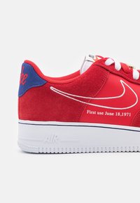 Červené tenisky vyrobené z semiše a síťoviny, bílé tkaničky a bílá podrážka. Obsahují modré akcenty a text uvádějící "První použití 18. června 1971."