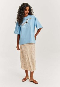 Světlě modré oversized tričko s grafickým potiskem a bílým textem, zkombinované s béžovou midi sukni s jemným opakujícím se vzorem. Hnědé ploché sandály.