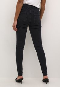 Jeans skinny taille haute en denim foncé, avec deux poches arrière et une texture lisse. Associés à des talons noirs ouverts.