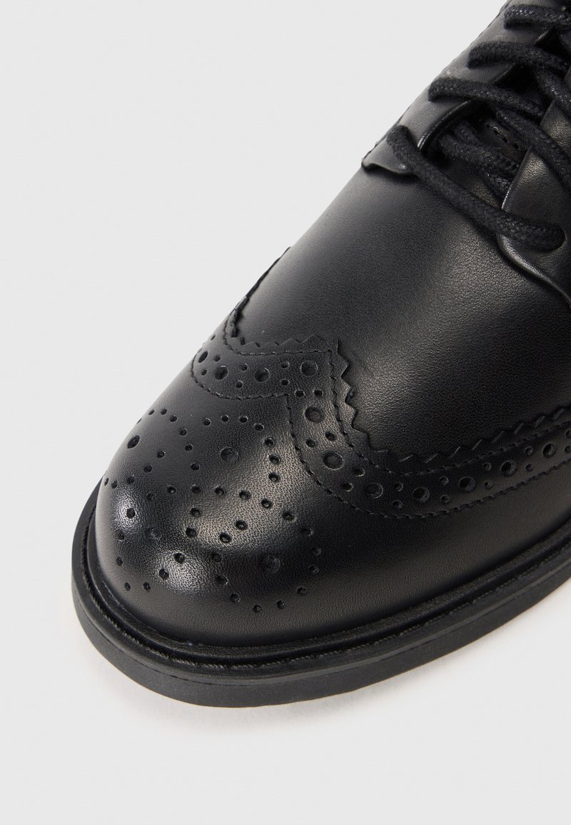 Zapato de vestir de cuero negro con punta redonda, intrincados detalles de brogue y cordones negros. Textura suave con un brillo sutil.