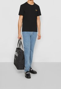 Svart bomull t-shirt med kort ärm och en liten gul logotyp, ljusblå denimjeans, svart väska och svarta slip-on skor med vit text.