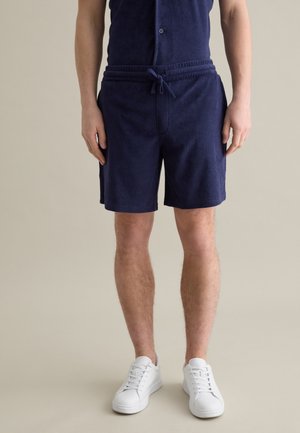 Pantalones cortos de toalla de color azul marino con cinturilla elástica y cordón. Presentan bolsillos laterales y un corte relajado. Llevados con zapatillas blancas.