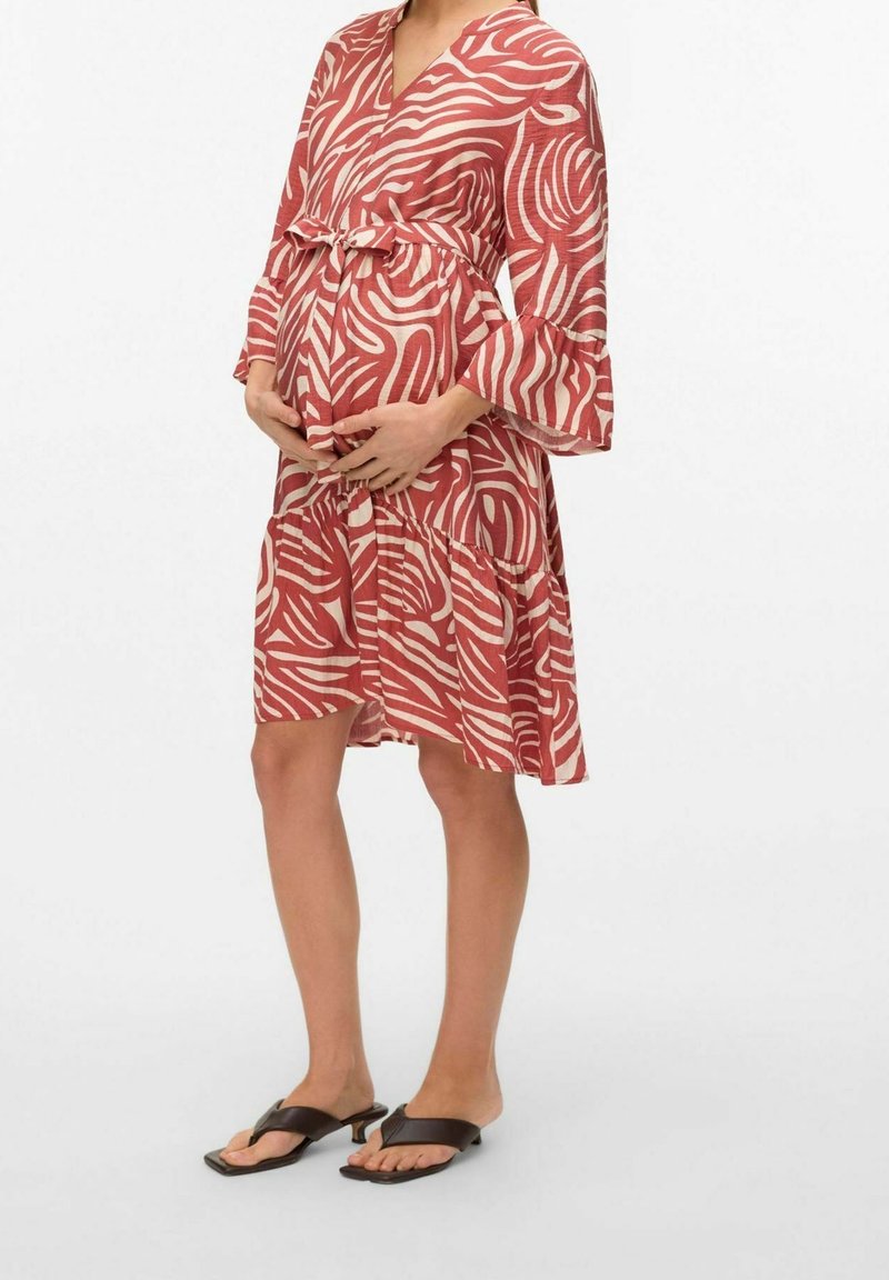 Femme enceinte portant une robe mi-longue à motifs rouges et blancs, tenant son ventre et portant des sandales à petits talons noires.