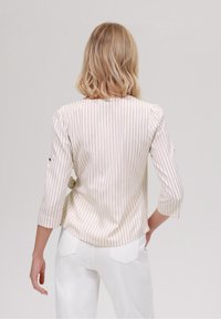 Pruhovaná blůza v béžové a smetanové tkanině s kulatým výstřihem, tříčtvrtečními rukávy a volným střihem. Pohled ze zadu ukazuje jemný detail nabírání.