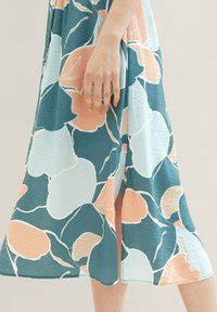 Robe midi fleurie avec un motif teal, pêche et blanc, en tissu texturé et fente sur le côté. Main reposant sur l'ourlet.