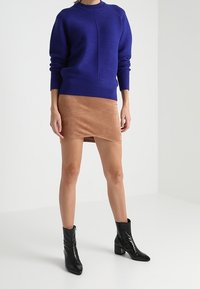 Pull en maille côtelée bleu avec col rond et manches longues, associé à une jupe portefeuille en daim beige. Des bottines noires viennent compléter la tenue.