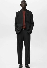 Wysoki mężczyzna w czarnym, oversize'owym garniturze z rdzawej swetrem, czarnym paskiem i czarnych butach wizytowych, stojący z rękami w kieszeniach.