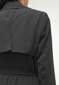 Czarny trench coat z wyraźnie zarysowanym kołnierzem, detalem w tylnej części oraz gładką teksturą i paskiem w talii.