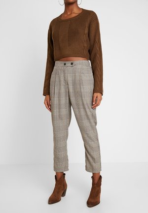 Femme portant un pull court marron en maille torsadée, un pantalon à carreaux taille haute avec boutons devant, et des bottines en daim marron.