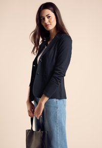 Schwarze Jersey-Blazer mit zwei Fronttaschen, Ein-Knopf-Verschluss und Reverskragen, kombiniert mit hellblauen weit geschnittenen Jeans und einer schwarzen Tragetasche.