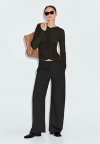 Maglietta nera a maniche lunghe con dettaglio arricciato, abbinata a pantaloni neri a gamba larga. Una borsa a tracolla marrone e occhiali da sole scuri completano l'outfit.