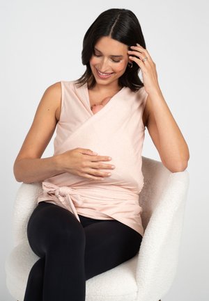Žena sedí na bielom kresle, nosí ružový zavinovací top a čierne legíny, drží a pozerá sa dole na bábätko zabalené v tope.