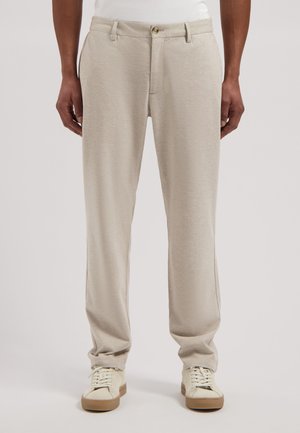 Beżowe spodnie o prostych nogawkach z przednimi kieszeniami, guzikiem i zamkiem błyskawicznym, noszone przez osobę w kremowych trampkach na jednolitym tle.