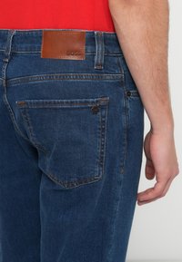 Tumesinised teksapüksid pruunist nahast plaastriga, millele on kirjaga "BOSS", oranži õmblusega ja tavapärase 5-tasku disainiga.