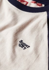 T-shirt en coton beige avec des accents marron marine sur le col et les manches, arborant un logo "Super Dry" brodé.