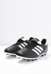 Des crampons de football en cuir noir avec des rayures blanches et un accent blanc autour de la cheville. Il dispose de lacets noirs et d'une semelle à picots moulés pour une meilleure traction.