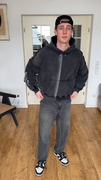 Nuori mies, jolla on musta huppari, taaksepäin käännetty lippalakki, harmaat farkut ja mustavalkoiset lenkkitossut, seisoo sisätiloissa puulattialla.