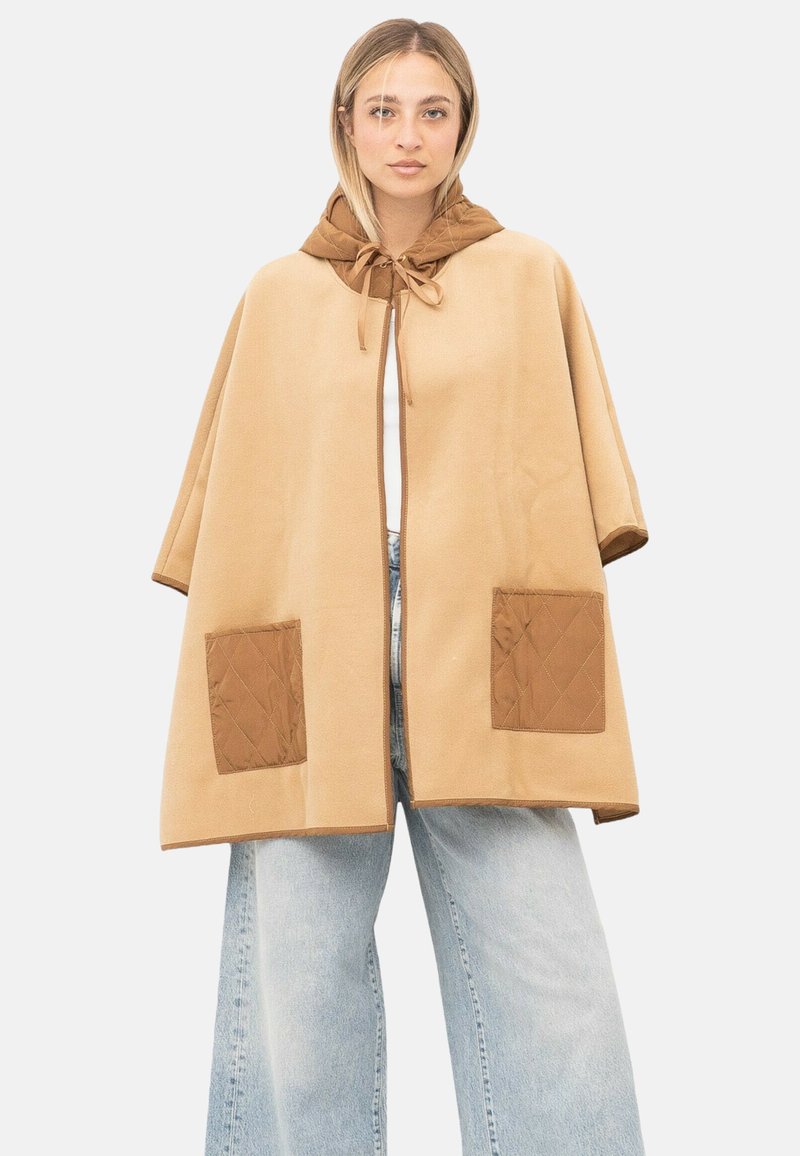 Vrouw draagt een beige cape met een bruin gewatteerde capuchon en zakken, gecombineerd met lichtblauwe wijde broekspijpen, tegen een effen achtergrond.