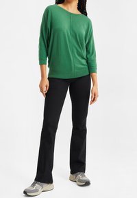 Grön lättstickad topp med trekvartslånga ärmar och vid halsringning, kombinerad med svarta rakbundna byxor och grå sneakers.