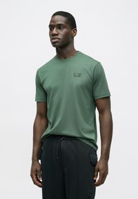 Olíva zöld rövid ujjú póló puha pamut anyagból, bal mellrészen finom fekete EA7 logóval nyomtatva.