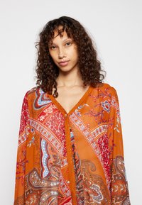 Blusa laranja fluida com intrincados padrões paisley em vermelho, azul e branco. Apresenta um decote em V e renda ao longo da frente.