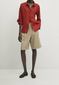 Chemise en lin rouge boutonnée avec manches retroussées, associée à un short beige longueur genou et des chaussures noires tissées sans lacets. Design simple, sans motifs.