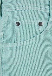 Tessuto in corduroy blu chiaro con righe verticali. Due bottoni metallici decorano una tasca laterale, aggiungendo un elemento di contrasto alla texture.