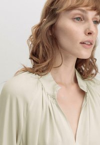 Hellgrüne, seidige Bluse mit tiefem V-Ausschnitt, gerafften Details am Kragen und locker sitzenden Ärmeln. Glatte Textur und minimalistisches Design.