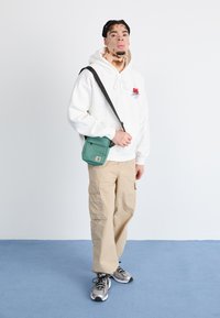 Hvit hettegenser med rød kirsebærgrafikk, beige cargo-bukser og en grønn crossbody-veske. Grå sneakers med svarte detaljer og snøring.