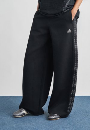 Pantaloni della tuta neri a gamba larga con tessuto testurizzato, caratterizzati da un logo Adidas bianco e dettagli metallici lungo i lati, abbinati a scarpe grigie.