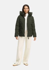 Quilted dark green puffer jacket with a hood, featuring a front zipper, side pockets, and a textured finish, worn over a white sweater.