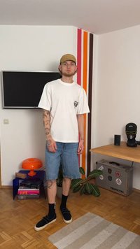 Valkoinen puuvillapaita, jossa on graafinen painatus, siniset farkkushortsit, mustat slip-on-kengät ja mustat sukat. Suljetussa ympäristössä värikkäät seinäraitat.