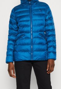 Blå uppblåsbar jacka med hög krage, dragkedja framtill och horisontella sömmar. Har sidofickor och en glänsande yta.