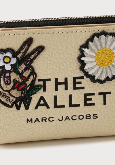 Μπεζ πορτοφόλι Marc Jacobs με κεντημένο χέρι σε σχήμα σήματος ειρήνης, επιθέματα μαργαρίτας, μαύρο φερμουάρ και μεγάλο κείμενο που γράφει "THE WALLET MARC JACOBS."