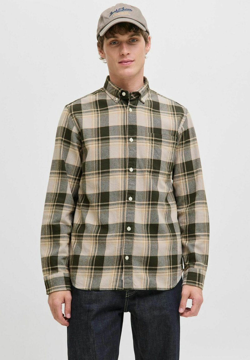 Camisa a cuadros verde y beige con mangas largas y cuello de botón; presenta pequeños botones y un corte relajado. Se lleva con pantalones vaqueros oscuros.