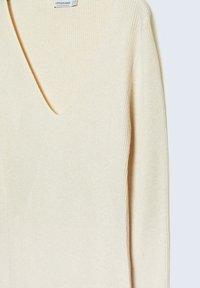 Pull en maille beige à col en V profond et manches longues. La texture du tissu semble douce et extensible, conçu pour une silhouette ajustée.