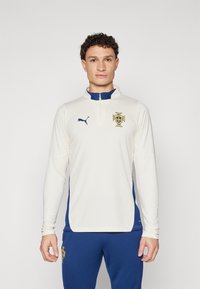 Kreemivärvi pikkade varrukatega sporditop, millel on tumesinine aktsent, kõrge krae ja Portugali embleem. Valmistatud tekstuursetest, hingavatest kangastest.