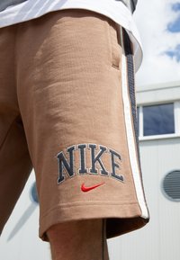 Bruna shorts med broderad "NIKE"-logotyp och röd swoosh. En del av en persons ben syns, med en byggnad och blå himmel i bakgrunden.