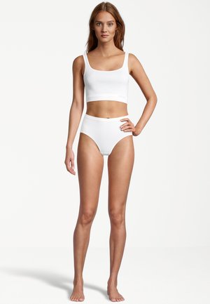 Weißes Baumwoll-Crop-Top mit breiten Trägern und hochgeschnittenen Slips, beide mit glatter Textur und schlichtem Design. Modell steht barfuß.