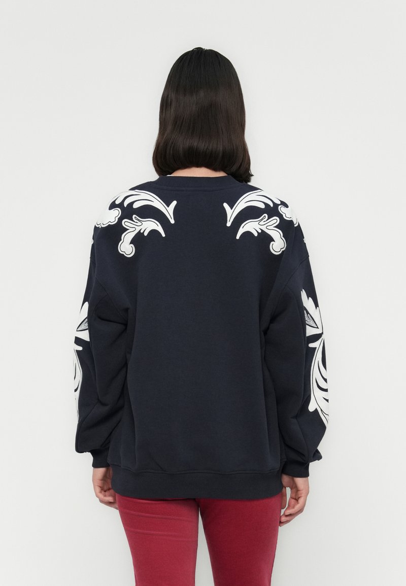 Navy sweatshirt met witte bloemenborduursel op de schouders en mouwen. Heeft een ronde halslijn en een relaxte pasvorm. Gepaard met rode broek.