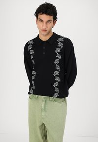 Zwart gebreid polo-shirt met bloemborduurwerk aan de voorkant, voorzien van een knopenlijst en lange mouwen, gecombineerd met lichtgroene broek.