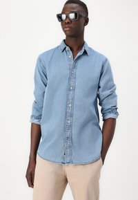 Cămașă din denim albastru deschis, cu guler fixat cu nasturi, buzunar pe piept și mâneci suflate, asortată cu pantaloni bej din țesătură și ochelari de soare negri.