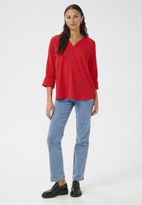 Rode blouse met lange mouwen, een V-hals en geplooide manchetten, gecombineerd met lichtblauwe spijkerbroek en zwarte instappers.