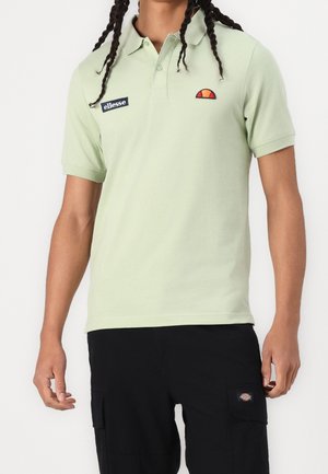 Mężczyzna w jasnooliwkowej koszulce polo marki ellesse i czarnych spodniach cargo z naszywkami, stojący na białym tle.
