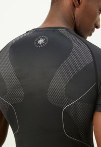 Gros plan sur un homme portant un tee-shirt de performance noir avec des panneaux gris à motifs et un petit logo circulaire dans le haut du dos.