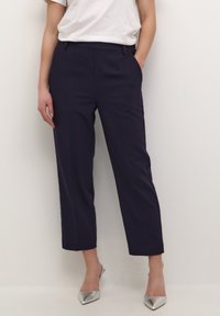 Pantalons taillés bleu marine avec une jambe droite, comportant des poches latérales, des passants de ceinture et un pli sur le devant. Portés avec des talons aiguilles argentés.