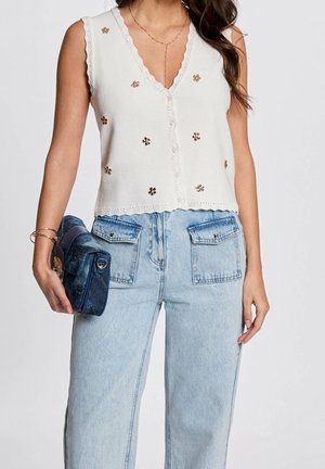 Kobieta w białej, bezrękawnikowej bluzce zapinanej na guziki z akcentami złotych kwiatów, jasnoniebieskich, wysokich dżinsach i trzymająca niebieską torebkę.