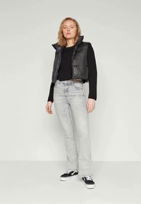 Svart polstret vest over en svart langermet skjorte, kombinert med lysegrå vide jeans og svart joggesko med hvite såler.