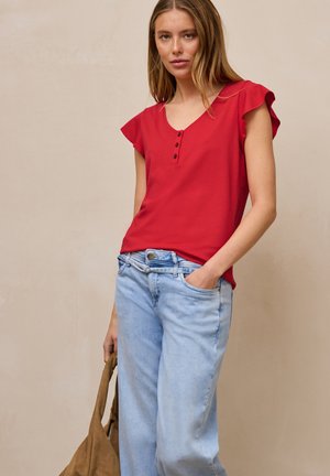 Vrouw draagt een rood kort mouwen blouse met knopen, lichtblauwe spijkerbroek, houdt een bruine tas vast, staat tegen een effen beige achtergrond.